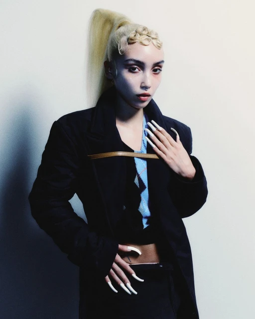 Adult female posing in a dark coat, blue top, and black skirt in front of a neutral background with a high blond ponytail hairstyle.