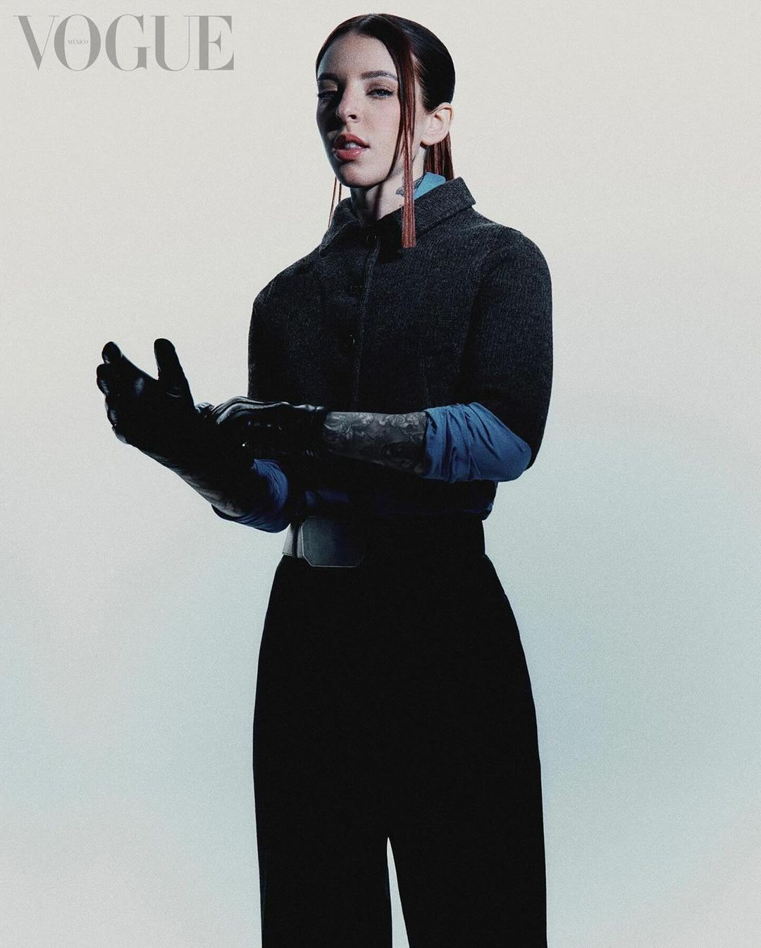 Fashion editorial shot of a model in black pants, a dark grey jacket with a blue shirt underneath, and black gloves, posing indoors.