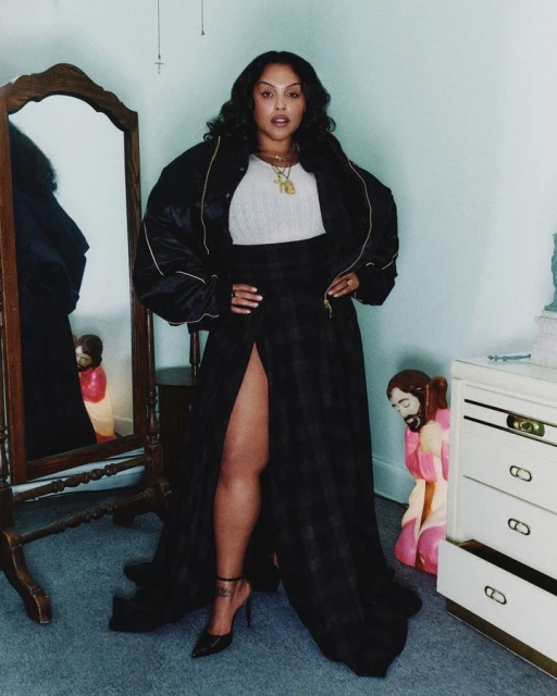 Adult woman in a black bomber jacket and plaid split maxi skirt, posing indoors with a mirror and a nightstand in the background.