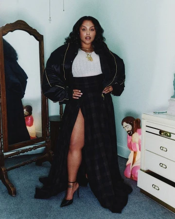 Adult woman in a black bomber jacket and plaid split maxi skirt, posing indoors with a mirror and a nightstand in the background.