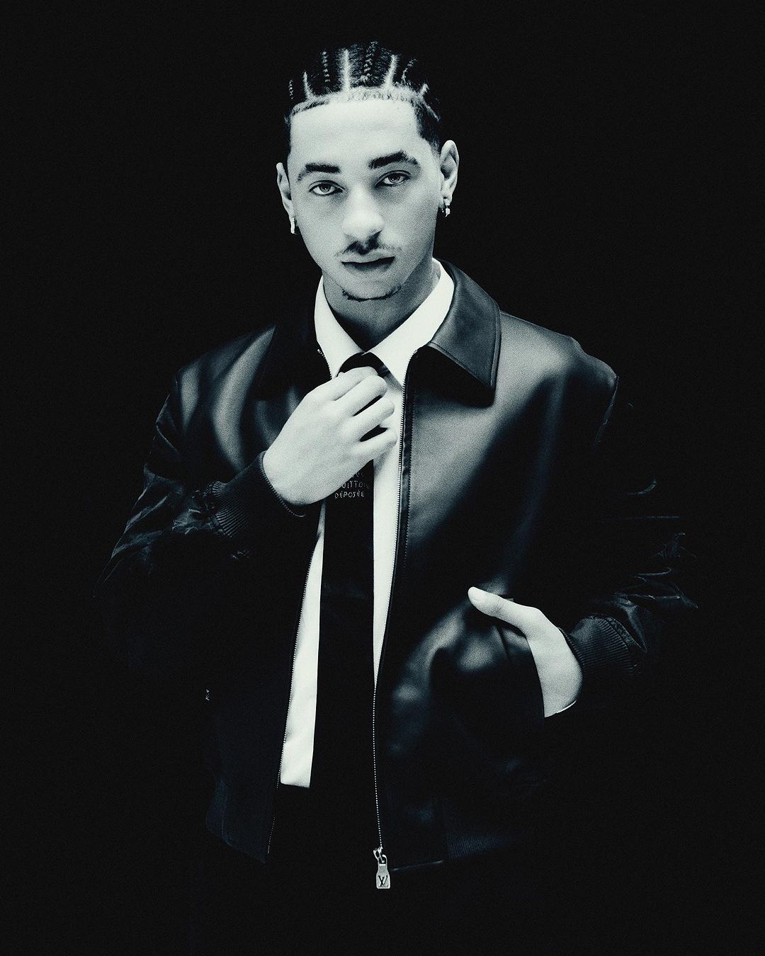 Monochromatic portrait of a stylish young adult male in a leather jacket adjusting his tie against a dark, high-contrast background.