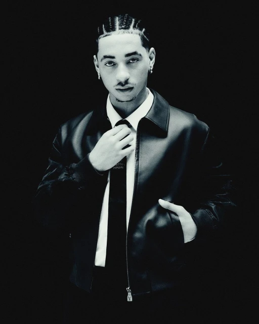 Monochromatic portrait of a stylish young adult male in a leather jacket adjusting his tie against a dark, high-contrast background.