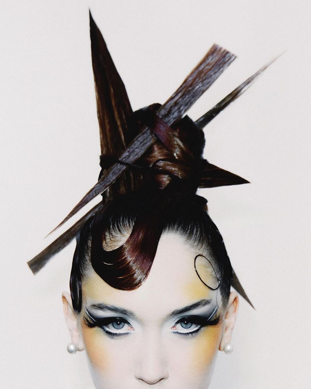 Close-up portrait of a woman with sculptural avant-garde brown hair and dramatic editorial makeup against a plain white background.