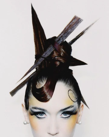 Close-up portrait of a woman with sculptural avant-garde brown hair and dramatic editorial makeup against a plain white background.