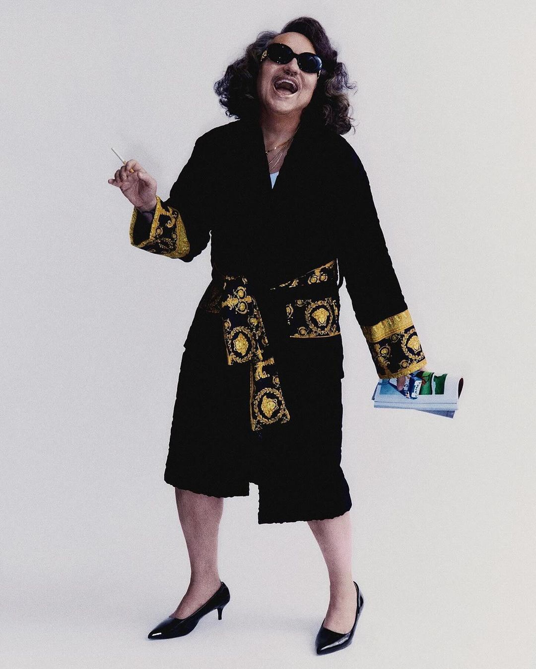 An adult in a black robe, sunglasses, and heels poses with a cigarette and magazines against a white background in a studio setting.