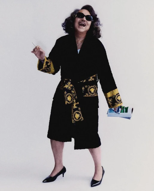 An adult in a black robe, sunglasses, and heels poses with a cigarette and magazines against a white background in a studio setting.