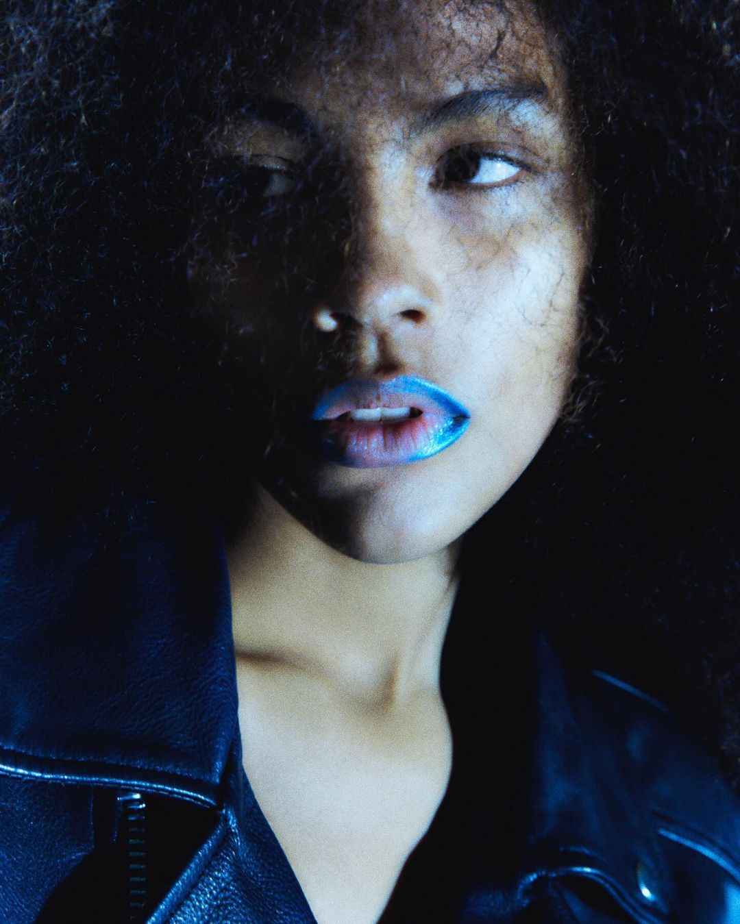 Close-up portrait of a Black woman with dark curly hair and blue lipstick, wearing a black leather jacket against a dark background.