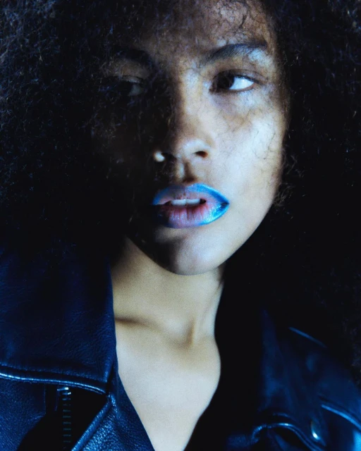 Close-up portrait of a Black woman with dark curly hair and blue lipstick, wearing a black leather jacket against a dark background.