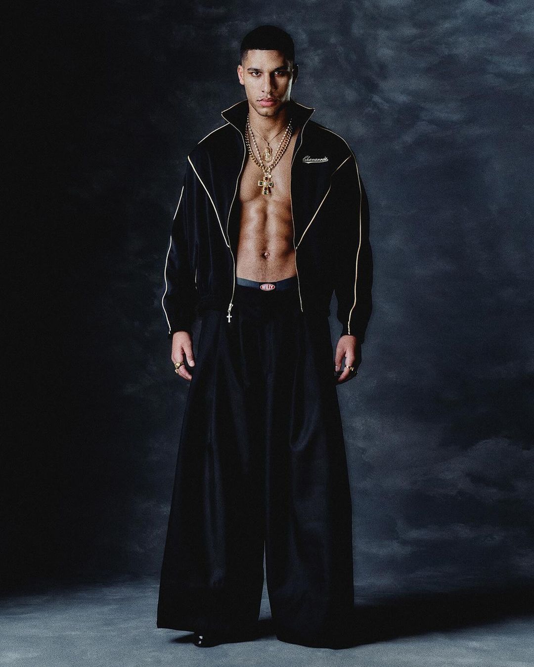 Studio shot of a young adult male in an open black tracksuit jacket, gold chains, and voluminous black trousers against a dark background.