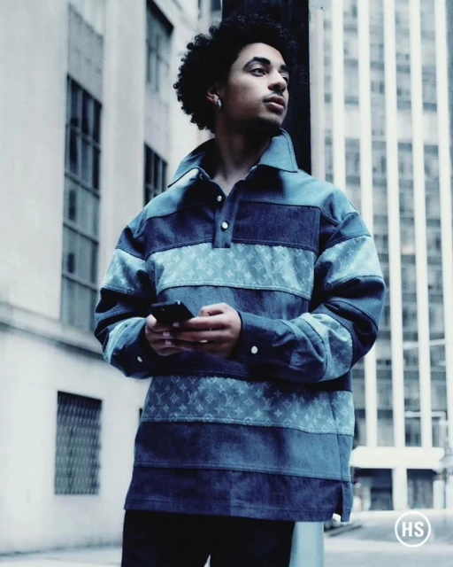 Young adult male with afro stands in an urban setting wearing a blue patterned jacket, holding a phone against city buildings.