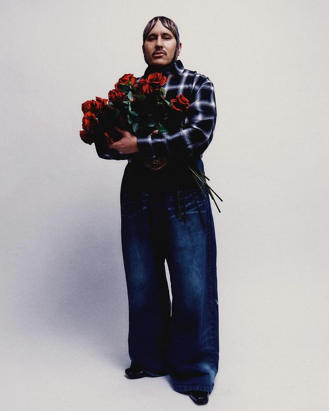 Adult male holds a bouquet of red roses, wearing an open black and white plaid shirt and wide leg blue jeans against a neutral backdrop.