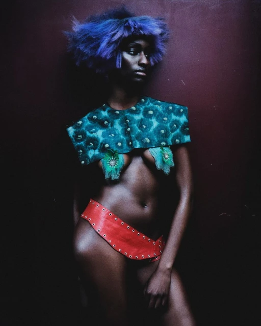 Striking portrait of a Black woman with creative teal and red fashion. She wears a blue wig and bold makeup in a dark studio setting.