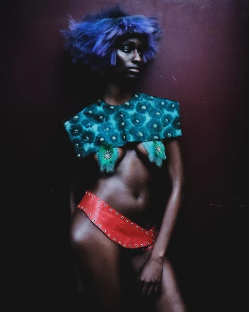 Striking portrait of a Black woman with creative teal and red fashion. She wears a blue wig and bold makeup in a dark studio setting.
