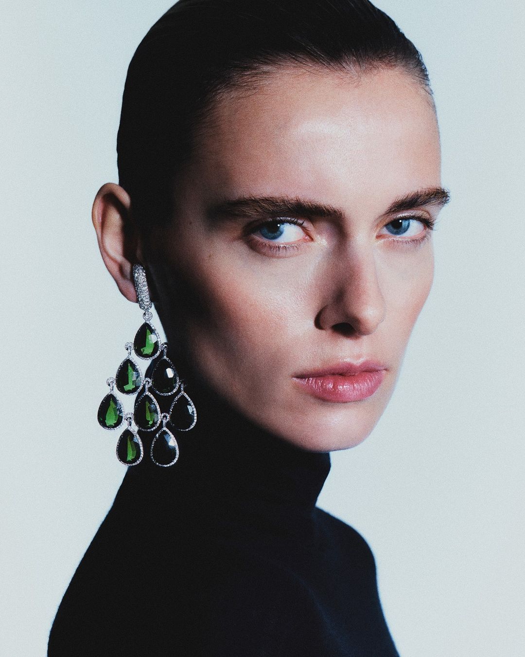 Close-up studio shot of a female model wearing dangling green gemstone earrings and a black turtleneck against a white backdrop.