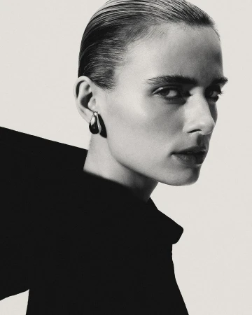 Monochrome portrait of a woman with slicked-back hair and statement earrings against a bright backdrop in a studio setting.