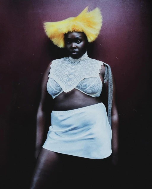 Dark-skinned woman poses in a white skirt, lace top, and yellow feathered headpiece against a solid burgundy backdrop in a studio setting.