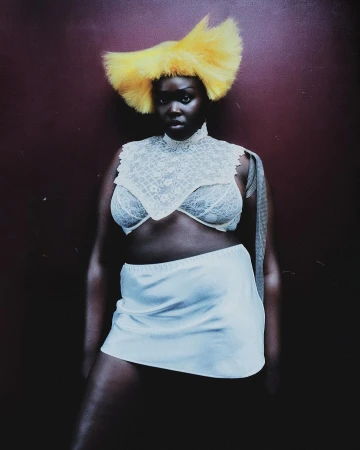 Dark-skinned woman poses in a white skirt, lace top, and yellow feathered headpiece against a solid burgundy backdrop in a studio setting.