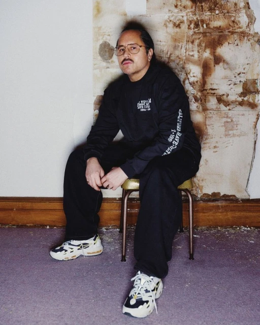 Adult male in glasses and black sweatsuit sits on a metal stool indoors against a wall with exposed plaster and wears white sneakers.