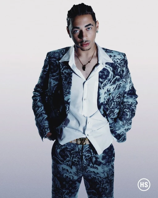 Man poses in a patterned suit, white button-down shirt, and gold necklace against a graduated white-to-grey backdrop in a studio shot.