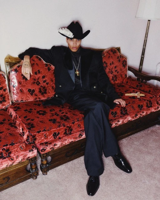 Man in black suit and cowboy hat lounges on red floral couch in an indoor setting, showcasing high fashion and classic elegance.