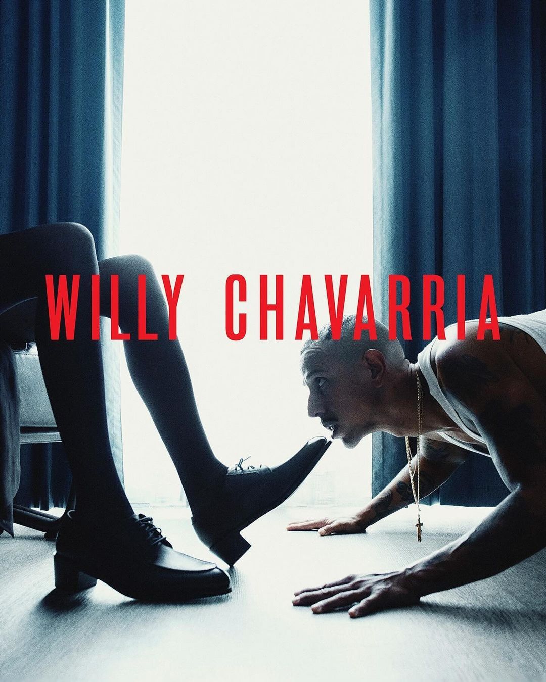 Man on floor with leg and shoe on head from Willy Chavarria fashion ad, shot in studio with curtain backdrop and Willy Chavarria logo.