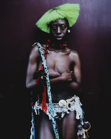Striking portrait of a woman with green hair and tribal-inspired accessories against a dark textured wall, expressive and culturally rich.
