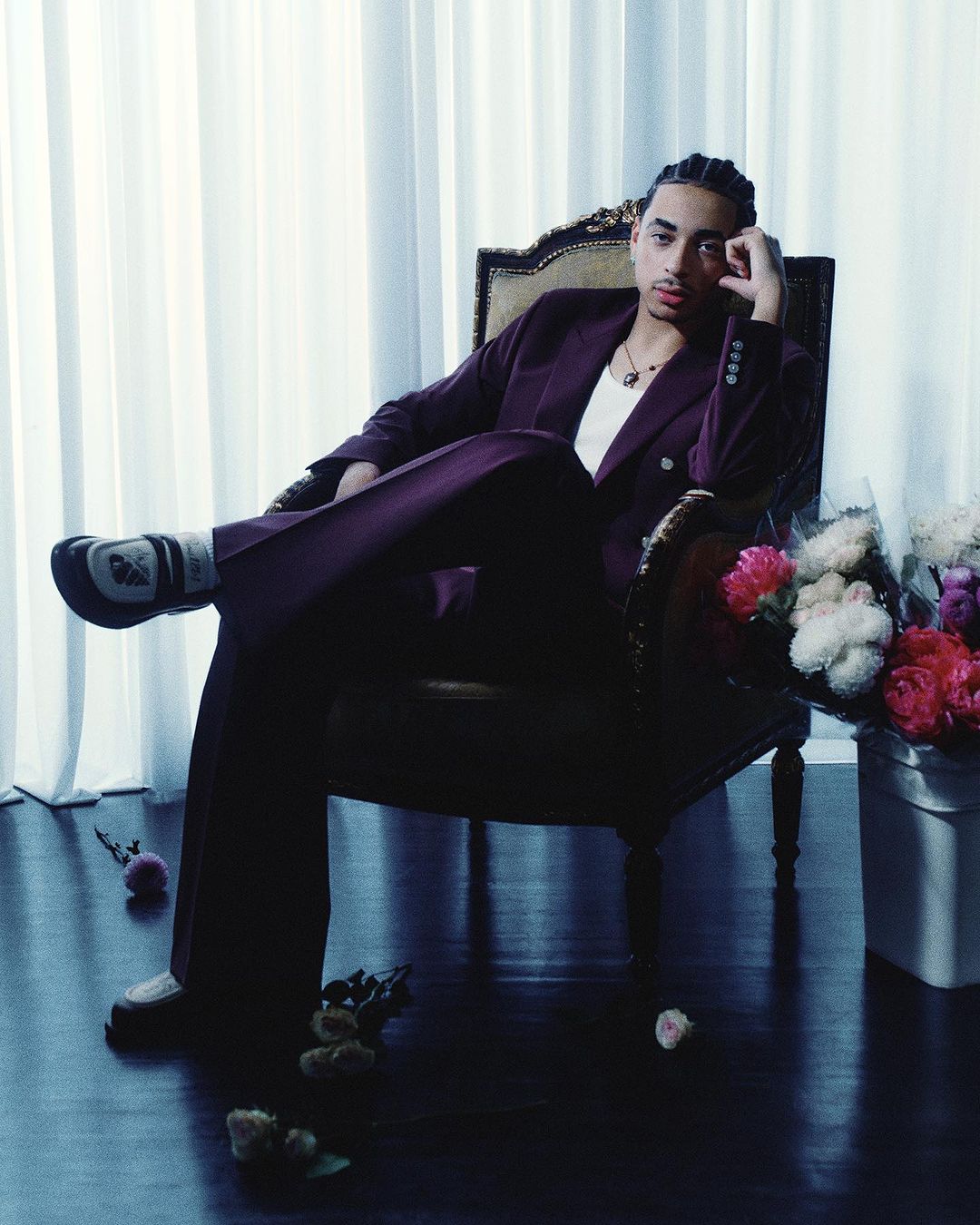 Man in a plum suit sits in an ornate chair with white flowers. He's resting his head on his hand and gazing into the distance.