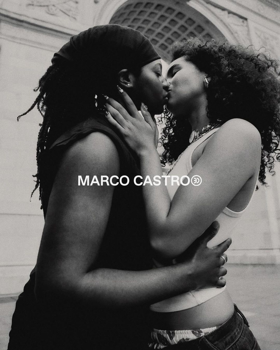 Two women are kissing in front of a stone archway in this black and white photograph. They're embracing and dressed in casual summer attire.