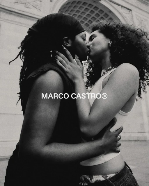 Two women are kissing in front of a stone archway in this black and white photograph. They're embracing and dressed in casual summer attire.