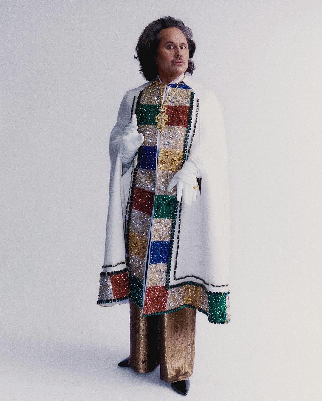 Full shot of a person with curly hair, wearing gold pants, black shoes, a white cape, and white gloves, standing against a white background.