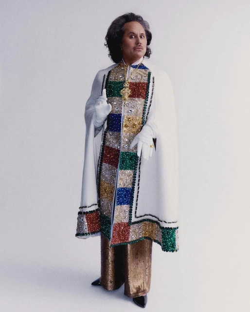 Full shot of a person with curly hair, wearing gold pants, black shoes, a white cape, and white gloves, standing against a white background.