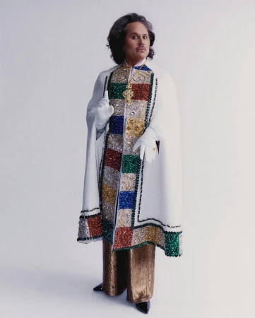 Full shot of a person with curly hair, wearing gold pants, black shoes, a white cape, and white gloves, standing against a white background.