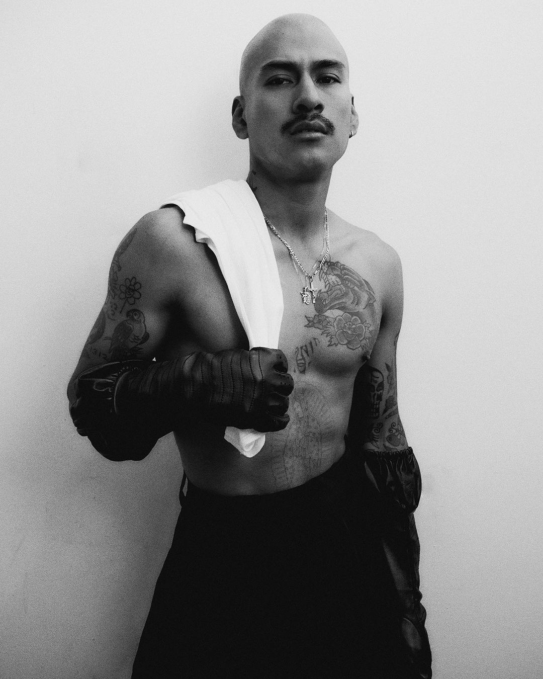 Black and white portrait of a shirtless man with shaved head, tattoos, and dark pants in front of a white wall.