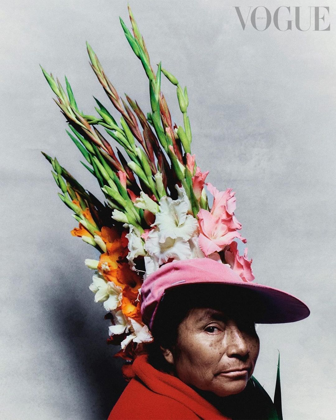 Portrait of a woman in a pink hat adorned with colorful gladiolus flowers against a light gray background with Vogue logo.