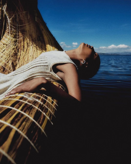 Woman in white dress rests on a straw boat on a lake under a blue sky. Calm water, bright sky, and natural textures dominate the image.