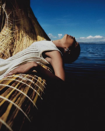 Woman in white dress rests on a straw boat on a lake under a blue sky. Calm water, bright sky, and natural textures dominate the image.