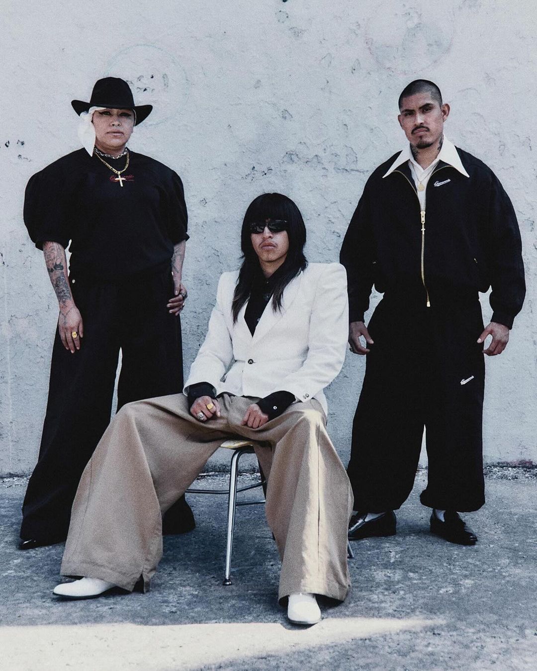 Three people pose against a textured white wall: a woman in black with a hat, a seated person in a white jacket, and a man in black.