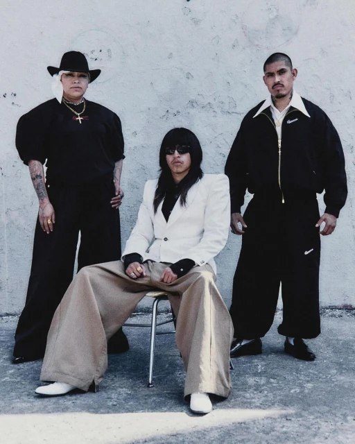 Three people pose against a textured white wall: a woman in black with a hat, a seated person in a white jacket, and a man in black.