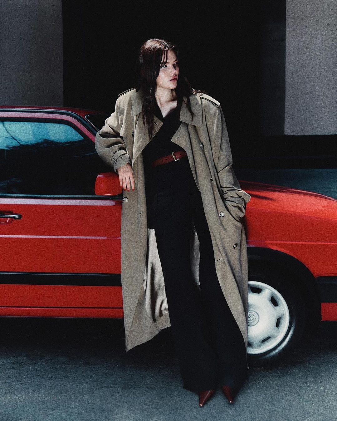 A woman in a trench coat poses next to a red car in an urban setting, showcasing fashion and automotive elements in soft lighting.