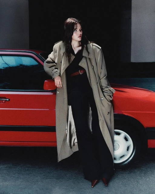 A woman in a trench coat poses next to a red car in an urban setting, showcasing fashion and automotive elements in soft lighting.