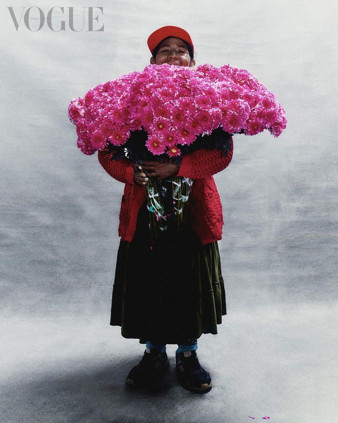 Woman smiles while holding a very large bouquet of bright pink flowers, posing in front of a grey background with the word VOGUE above.