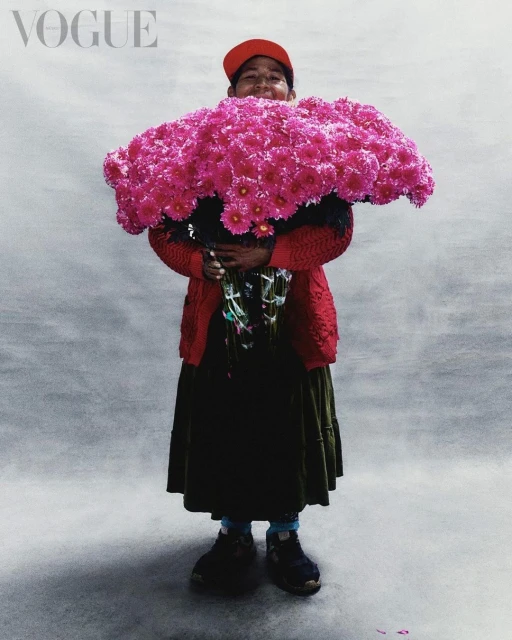 Woman smiles while holding a very large bouquet of bright pink flowers, posing in front of a grey background with the word VOGUE above.