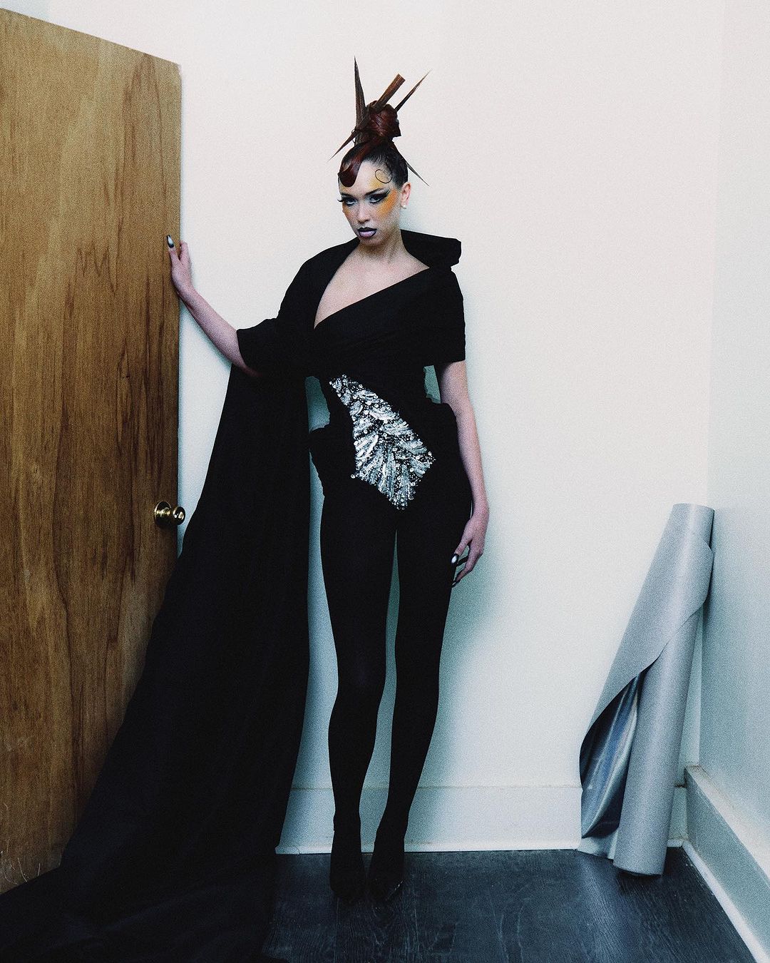 Woman with avant-garde hair and fashion stands near a door and silver-colored roll of material in an indoor corner setting.