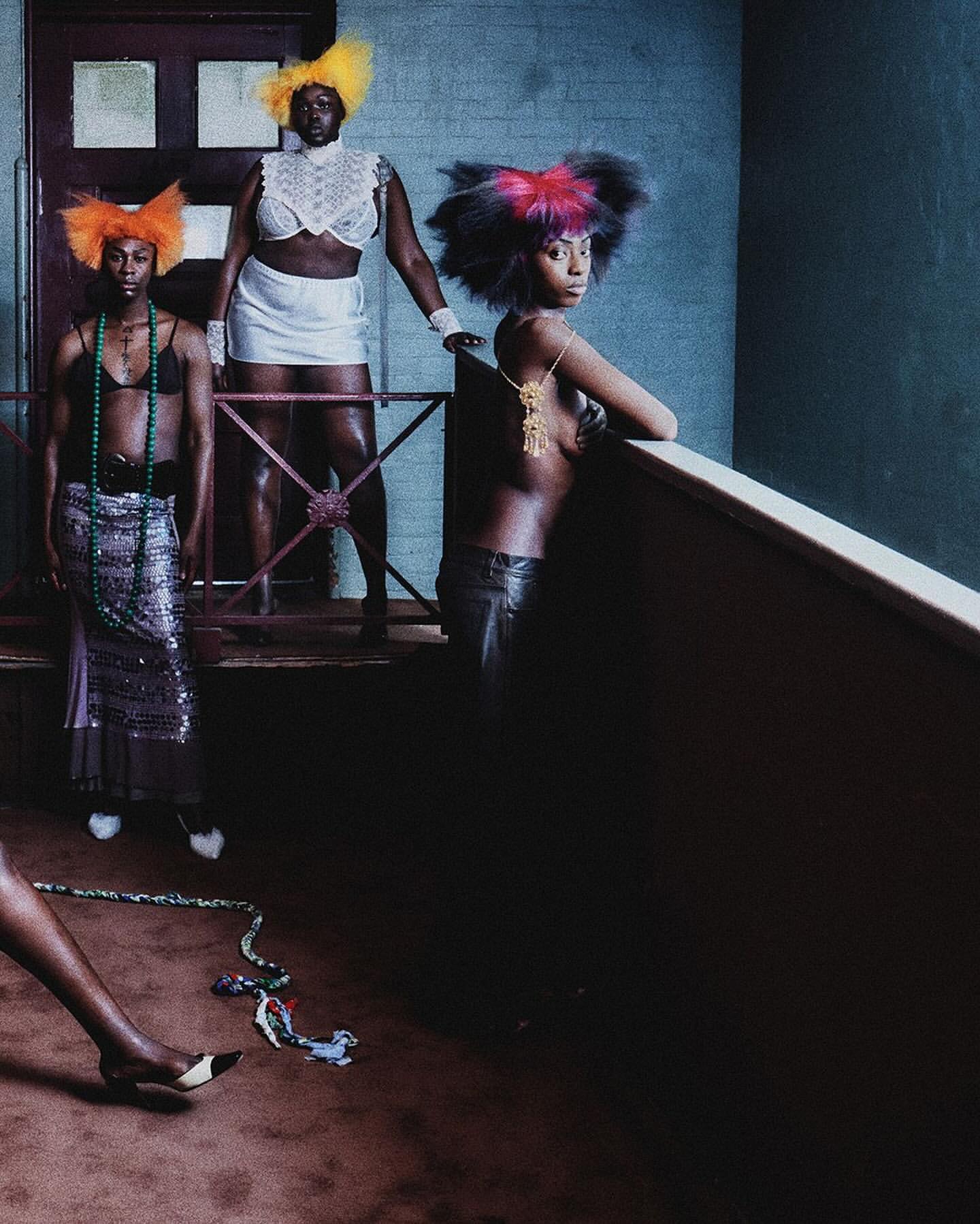 Three striking figures with colorful hair and unique outfits pose indoors against textured walls with artistic flair and creative expression.
