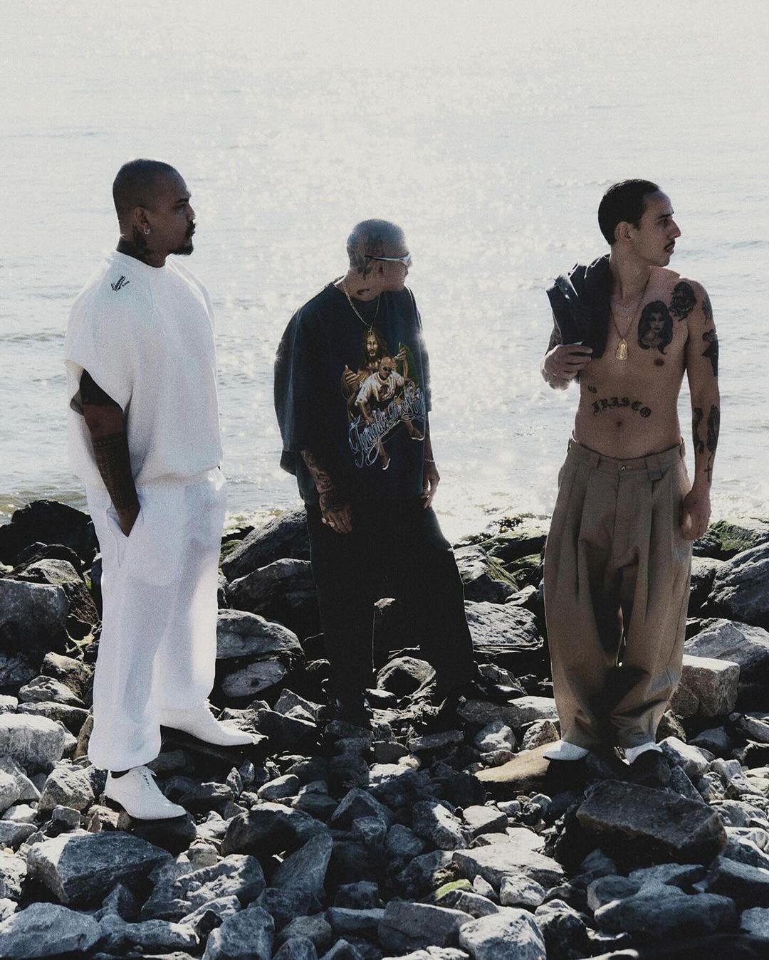 Three tattooed men stand on a rocky shore with the ocean in the background. Two wear shirts, one is shirtless with trousers.
