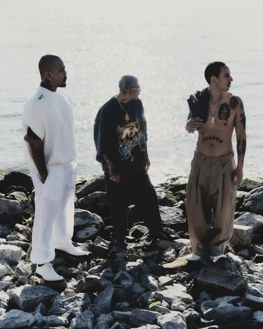 Three tattooed men stand on a rocky shore with the ocean in the background. Two wear shirts, one is shirtless with trousers.