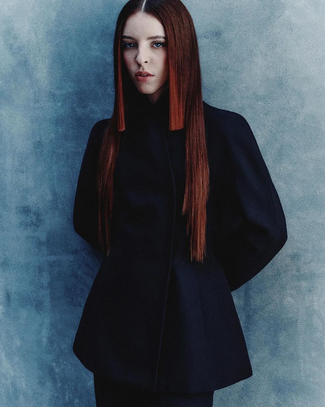 Portrait of a woman with long red hair wearing a dark structured jacket against a mottled blue-grey textured wall.