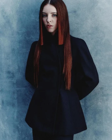 Portrait of a woman with long red hair wearing a dark structured jacket against a mottled blue-grey textured wall.