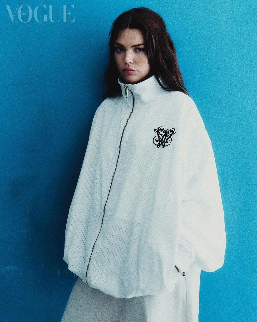 A model poses against a blue wall wearing a white jacket and pants in a studio shot for Vogue magazine cover; fashion editorial.