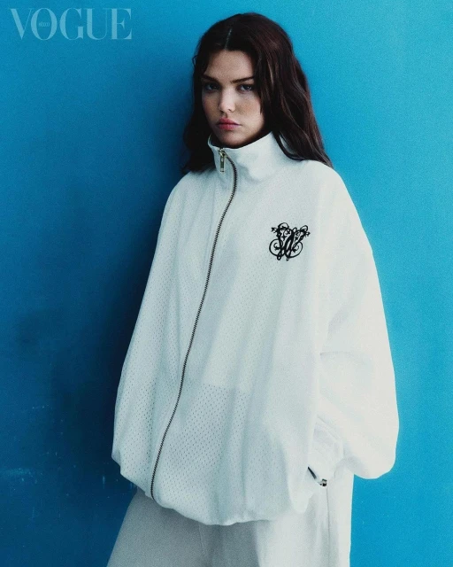 A model poses against a blue wall wearing a white jacket and pants in a studio shot for Vogue magazine cover; fashion editorial.